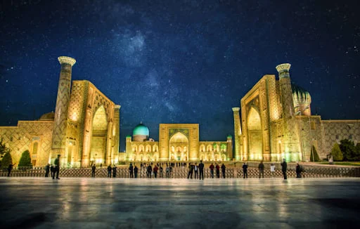 ثلاث واجهات مدارس في ساحة ريغستان بسمرقند، تضيء بواباتها المكسوّة بالقاشاني شمسُ المساء المنخفضة.