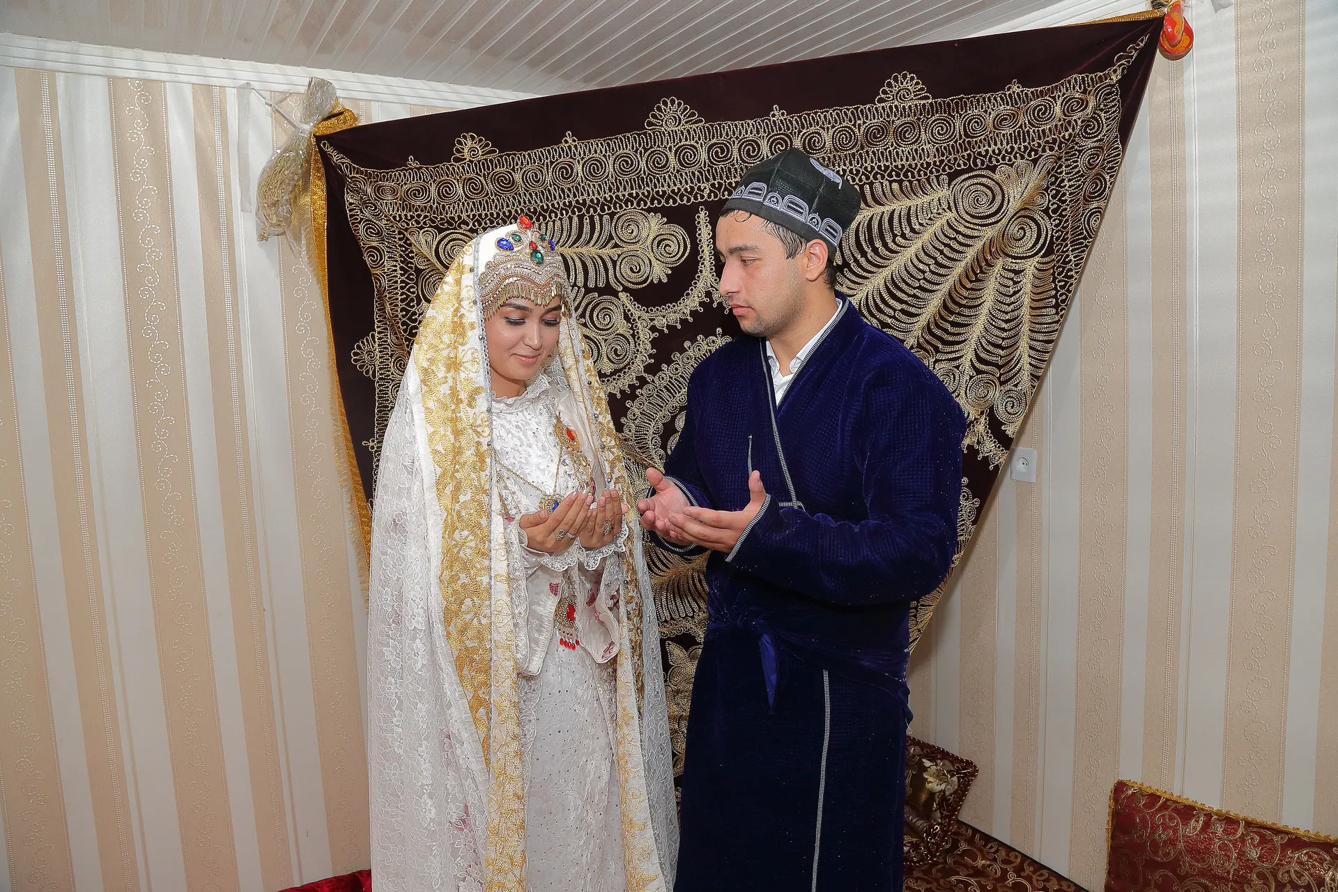 Bride and groom in traditional dress after the first day of their wedding, beneath an embroidered wall hanging.